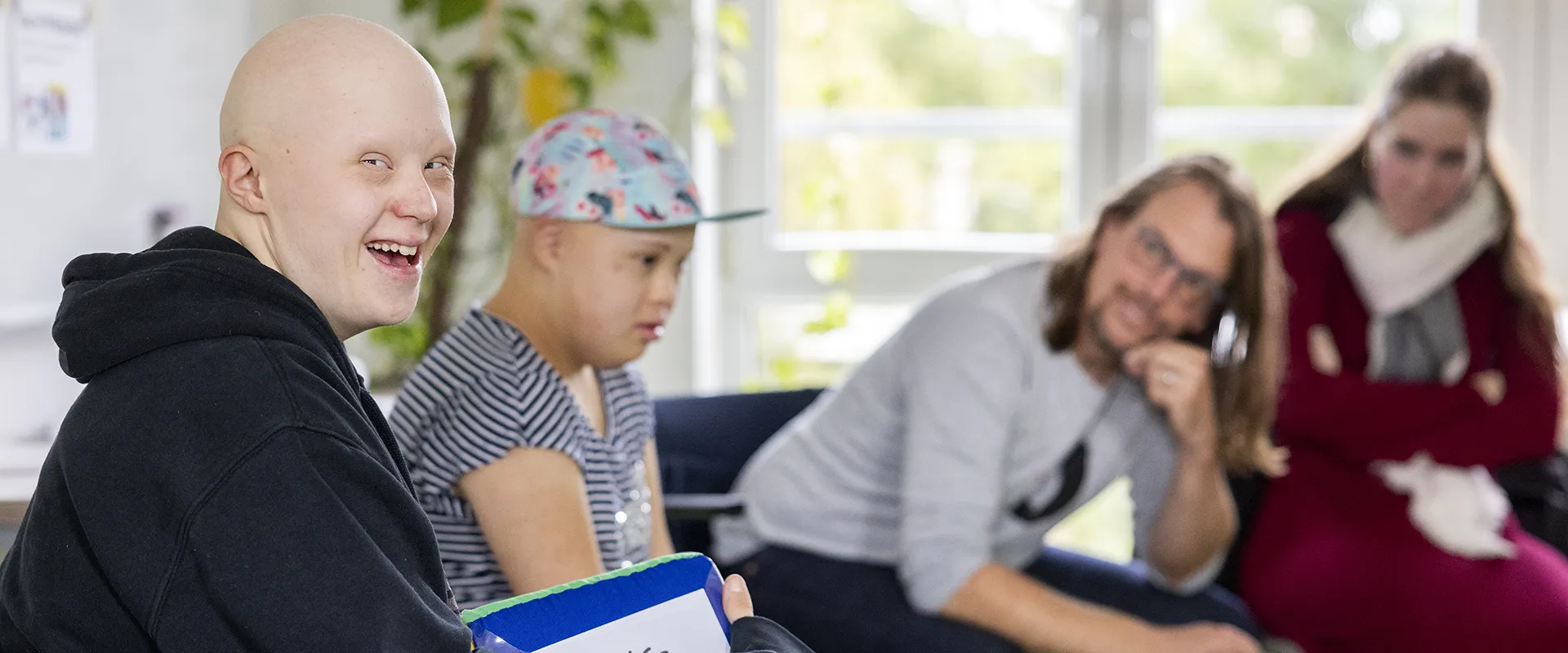 Eine Gruppe von vier Personen in einem Innenraum. Die Person ganz links schaut in die Kamera und lacht, die anderen drei sind unscharf im Hintergrund zu sehen.
